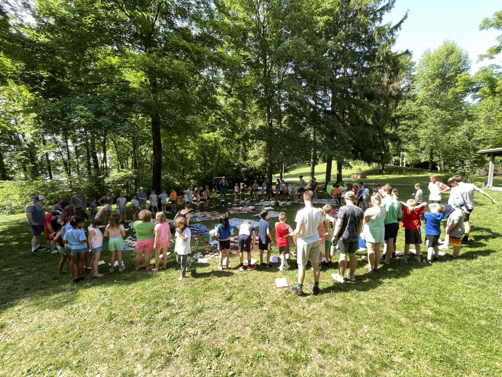 Campers and counselors standing in a wide circle on the grass among tall trees at Camp Friedenswald.