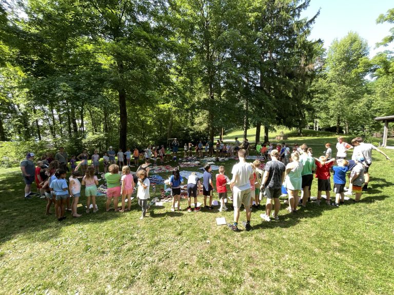 Campers and counselors standing in a wide circle on the grass among tall trees at Camp Friedenswald.