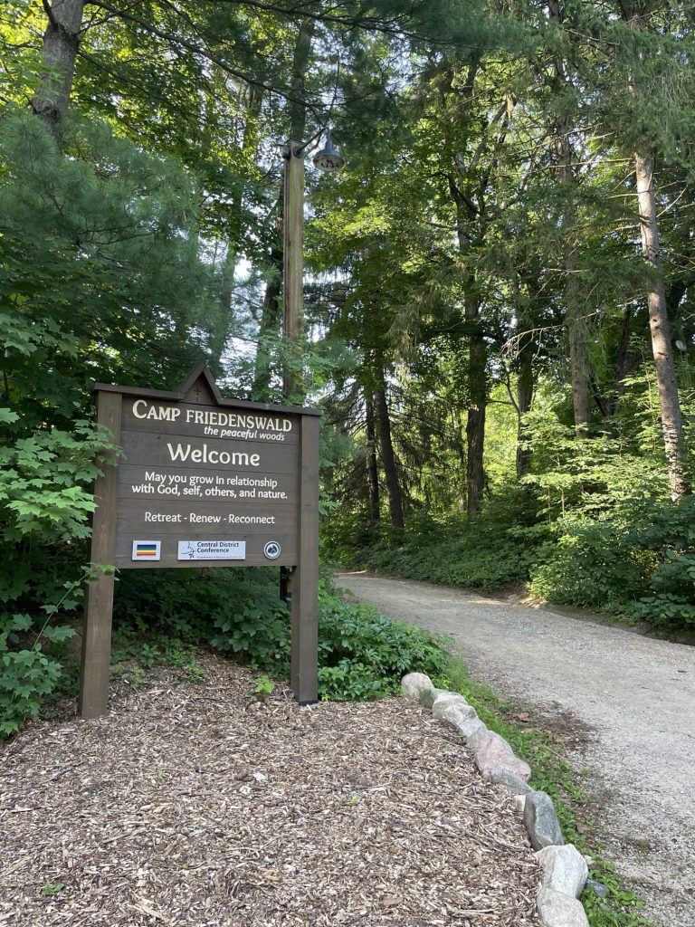 Wooden sign at the entrance to Camp Friedenswald reading Welcome — May you grow in relationship with God, self, others, and nature.