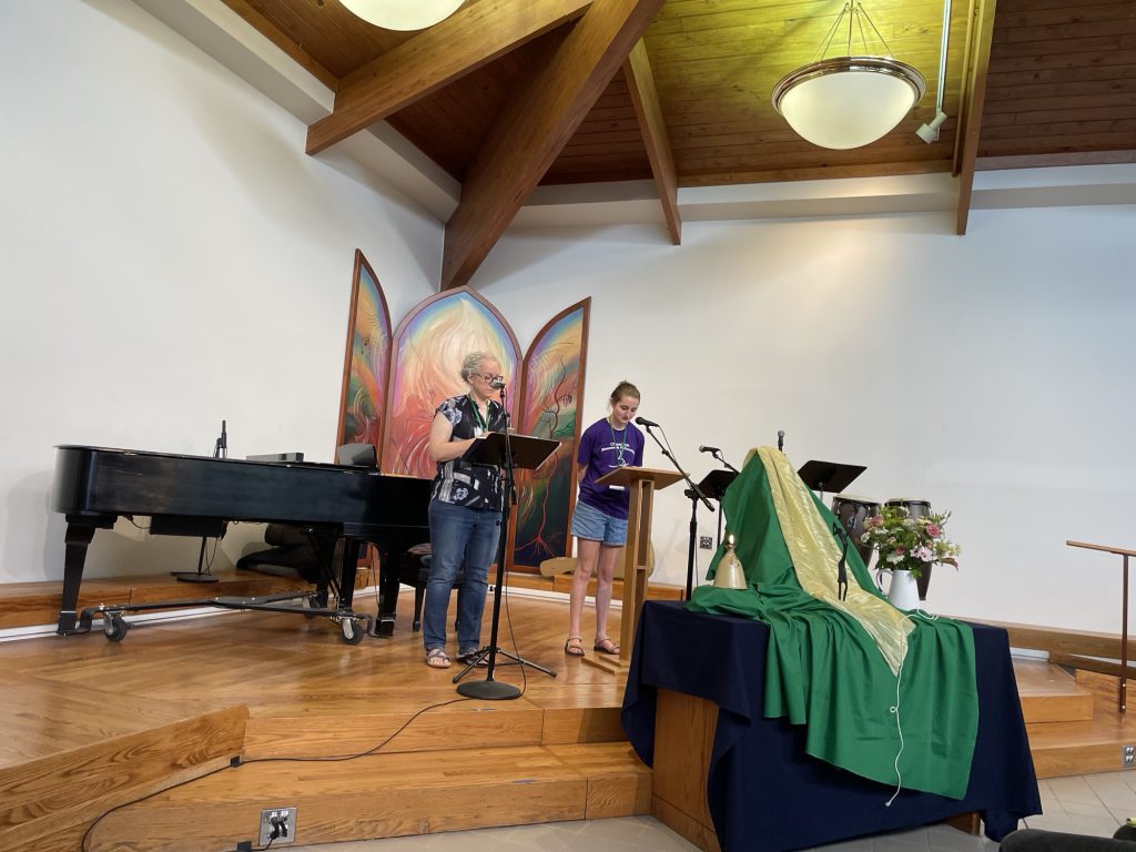 Two worship leaders reading from music stands beside a grand piano and a green-draped communion table.